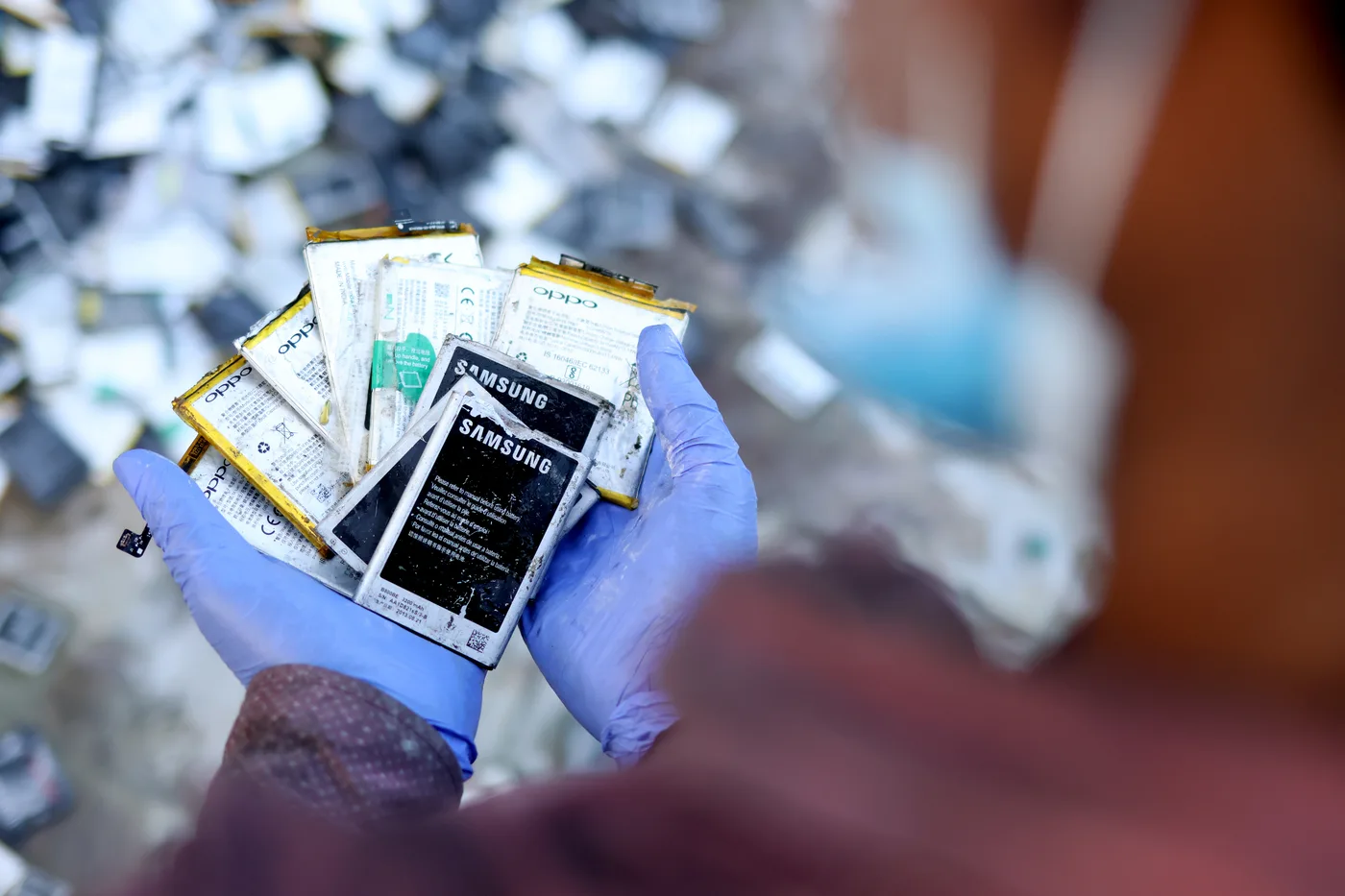 A gloved hand holding a fan of spent phone batteries over a large pile of collected e-waste batteries