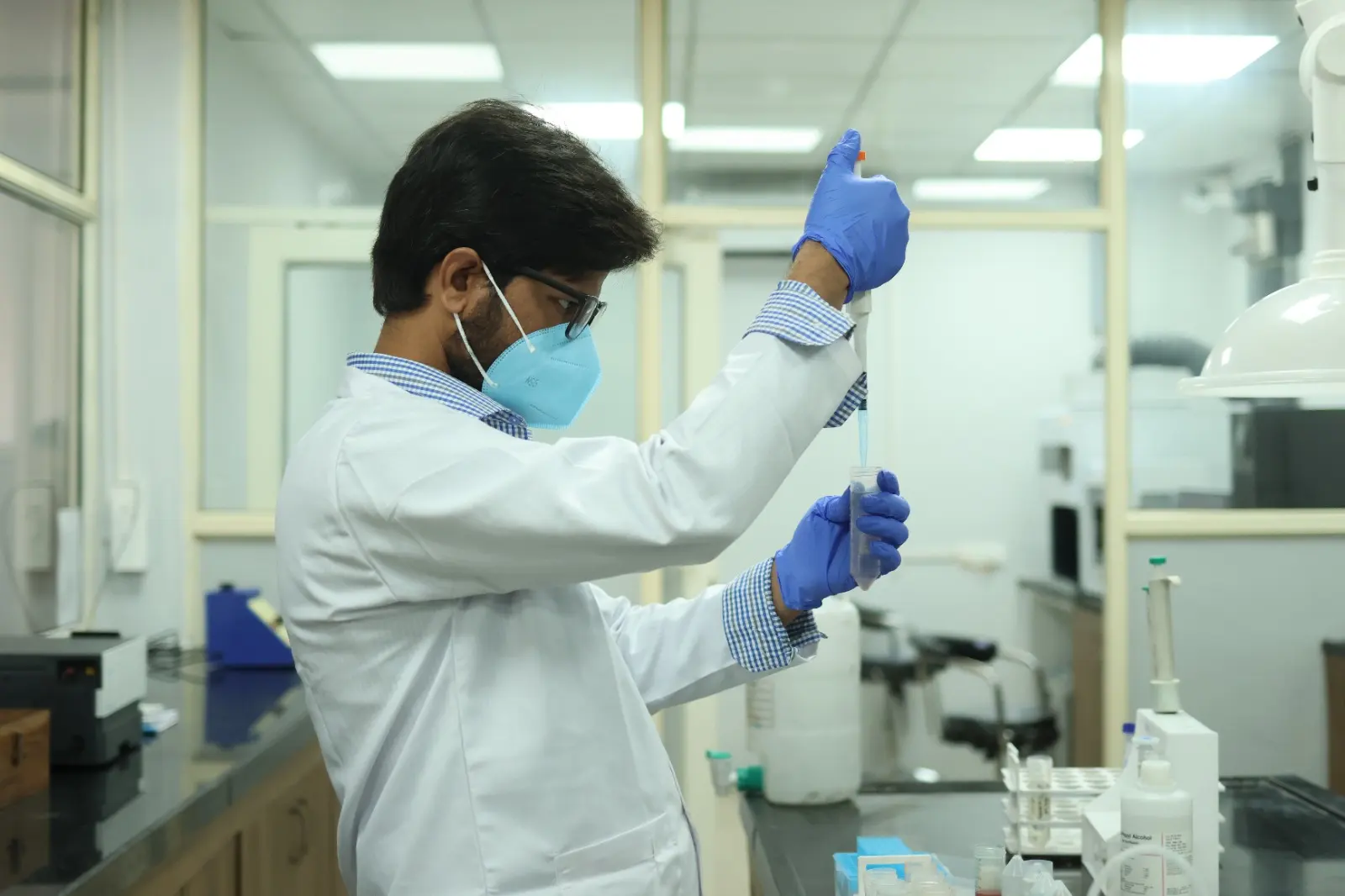 A technician in a white coat, mask and gloves holding a pipette up to the light