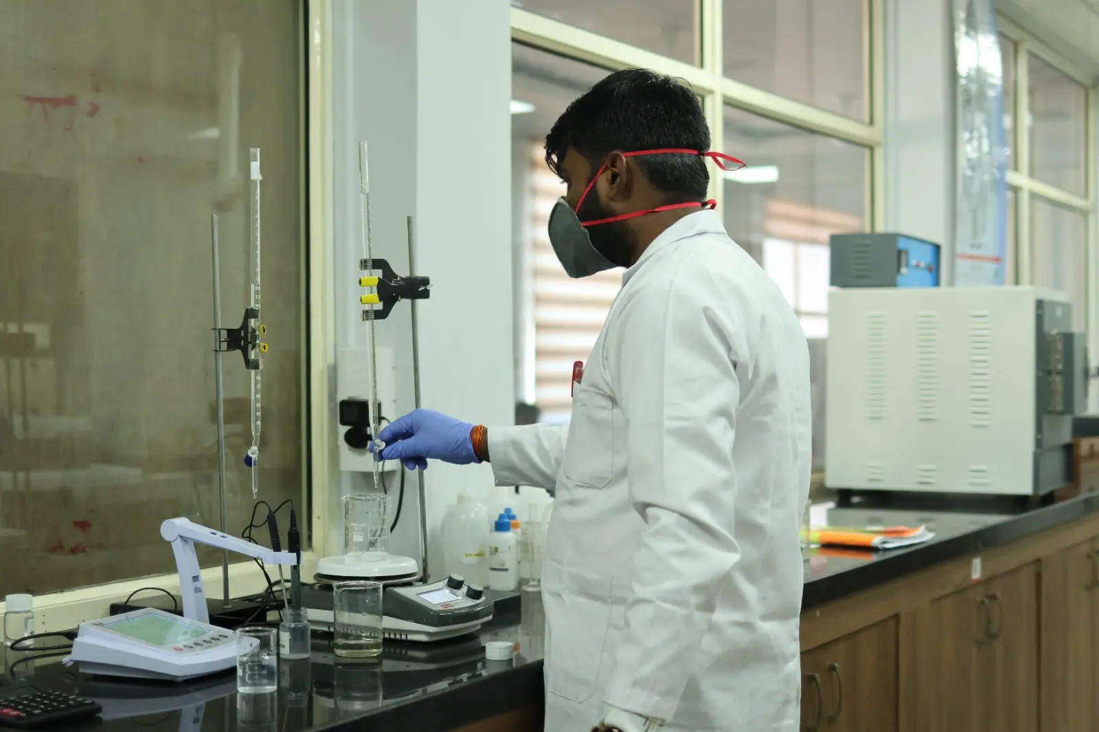 A technician in a white coat, mask and gloves working at a titration stand on a lab bench