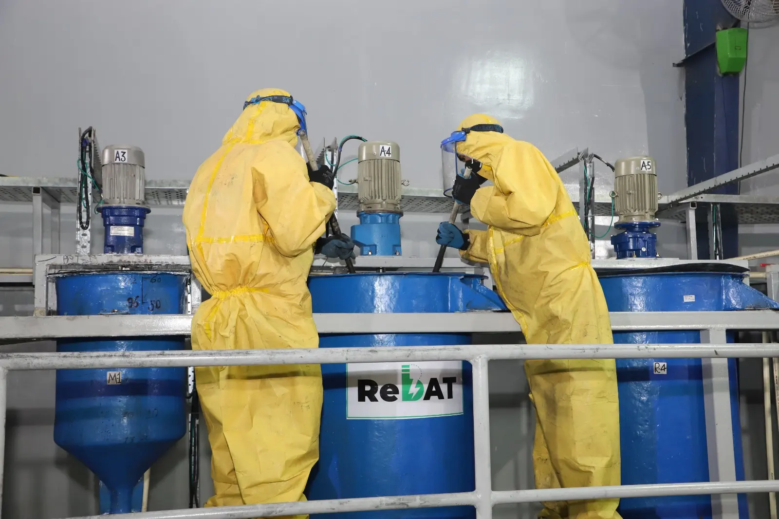 Two workers in yellow protective suits and face shields at ReBAT-branded blue tanks