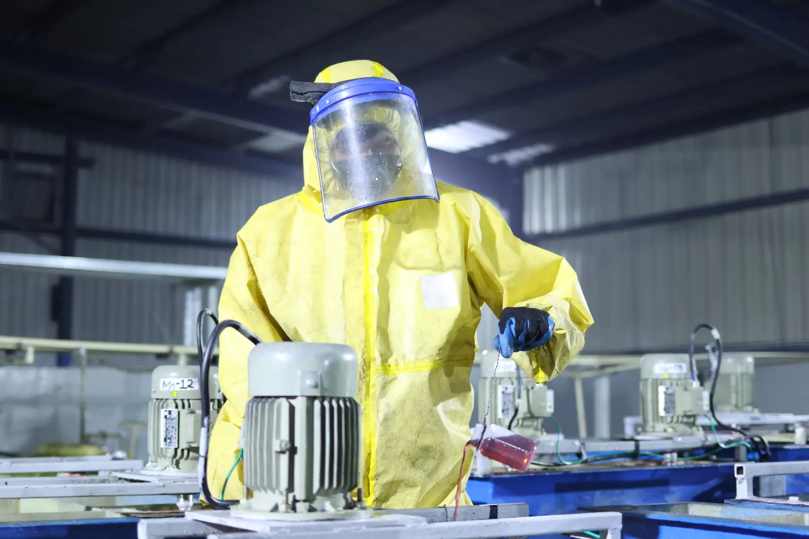 A worker in protective gear holding a small sample flask of pink liquid beside a motor